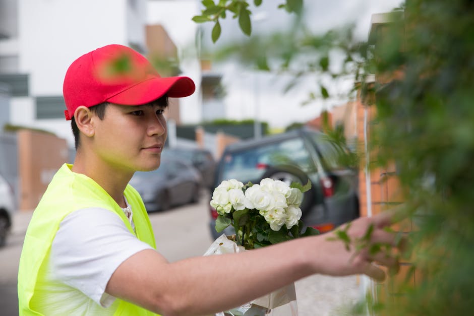 A young male florist wearing a red cap and a high-visibility yellow vest is placing a bouquet of white hydrangeas wrapped in paper and tied with a white ribbon against a brick wall. The arrangement fe