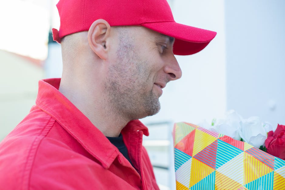 A delivery person wearing a red cap and matching red jacket, captured in profile, smiling as they hold a colorful, geometric-patterned gift bag filled with white and pink flowers, likely roses or simi
