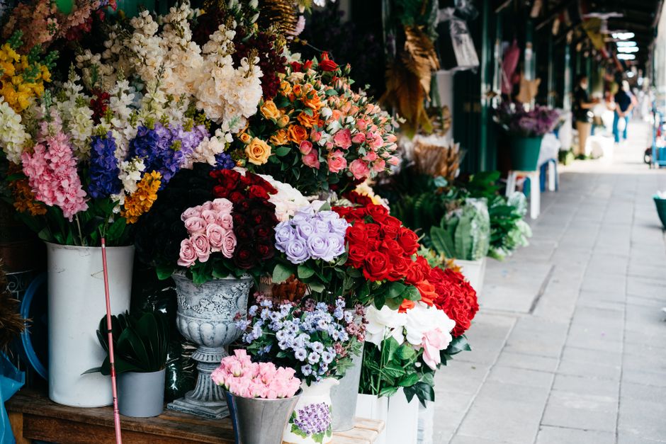 An outdoor display of vibrant floral arrangements and bouquets arranged in various ornate vases and buckets along a sidewalk. The flowers include roses, hydrangeas, and other mixed blossoms in shades