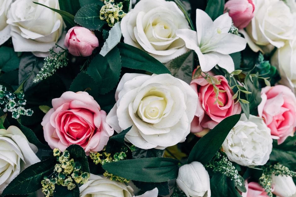 A close-up of a floral arrangement featuring predominantly white and pink roses, with some white lilies and small clusters of delicate white and green filler flowers. The bouquet is lush with dark gre