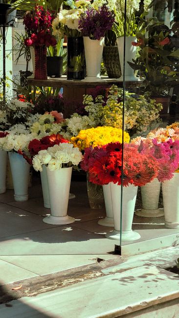 A display of various fresh floral arrangements in white ceramic and glass vases positioned on a glass shelf and on the ground outside a flower shop. The arrangements include a mix of white, yellow, re