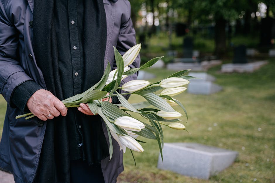 A person dressed in a dark jacket and shirt holding a bouquet of white lilies with green stems and leaves. The flowers are fresh and partially unopened, arranged in a natural style. The bouquet is gra
